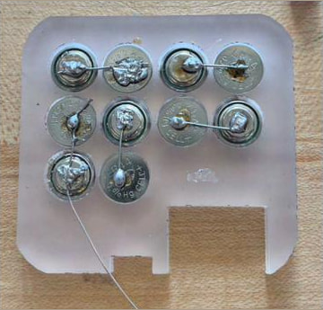 A clear acrylic plate carrying nine coin cells joined by solder links and wire