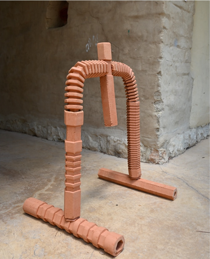 An assembled terracotta cooling screen standing on a concrete floor: ribbed hexagonal clay pipes bend into an arch over a straight base pipe