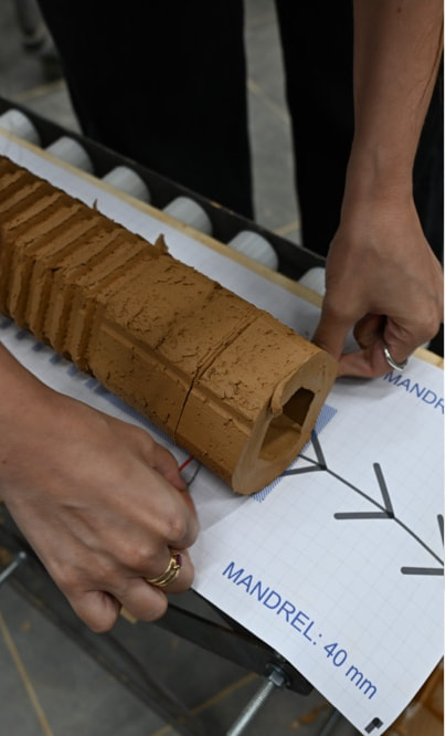Hands guide a ribbed clay pipe onto the printed paper blueprint, which is labelled with the mandrel size