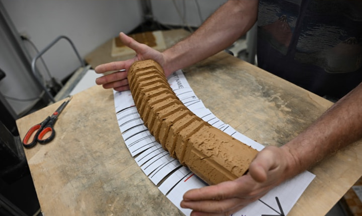 Two hands bend a ribbed clay pipe along the cut lines of a printed blueprint on a workbench