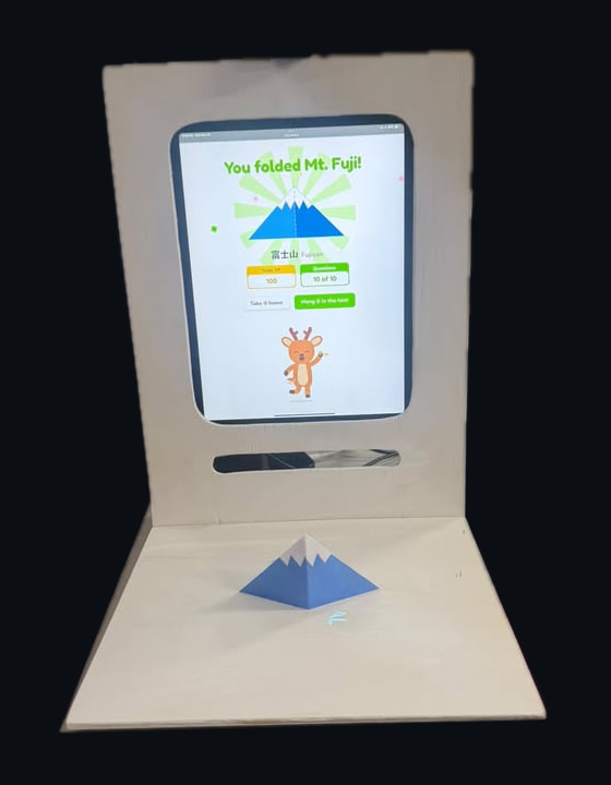 The Fold with Nara booth: a pale wooden stand holds an iPad showing “You folded Mt. Fuji!” above a folding tray, with a blue and white paper Mt. Fuji standing on it